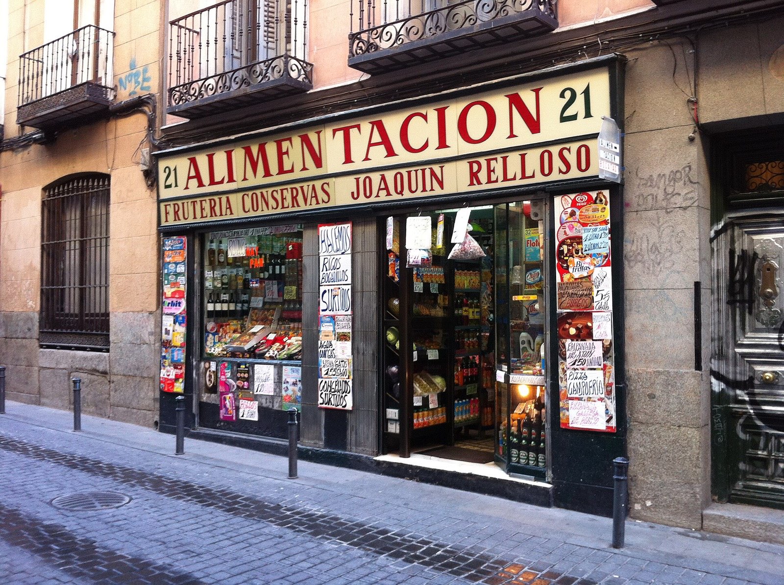 Fachada de una tienda de ultramarinos en una calle empedrada de Madrid, con el escaparate lleno de carteles